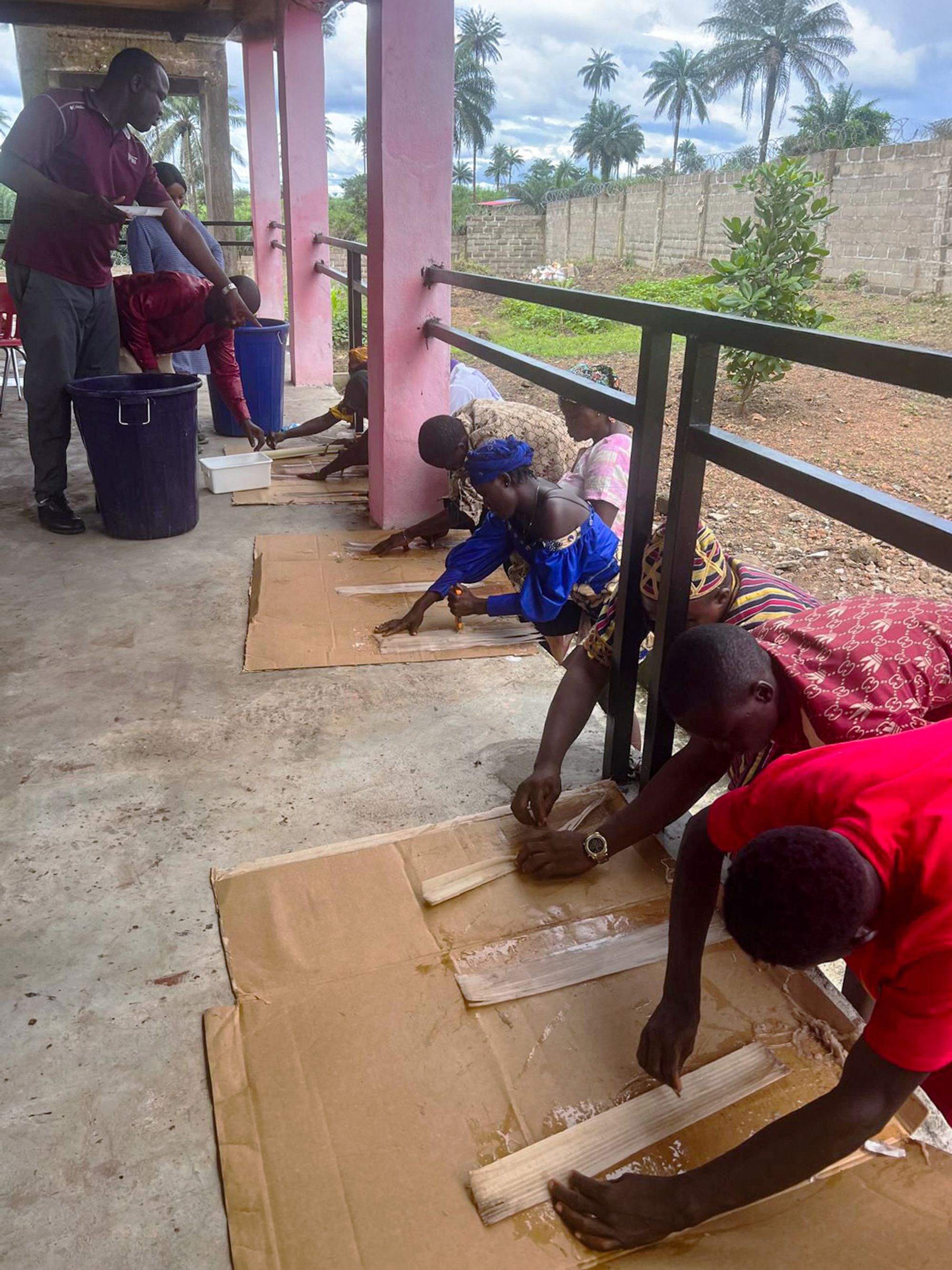 A group of people crouched on a concrete floor under a shaded structure, processing banana plant fibers laid out on large sheets of cardboard.